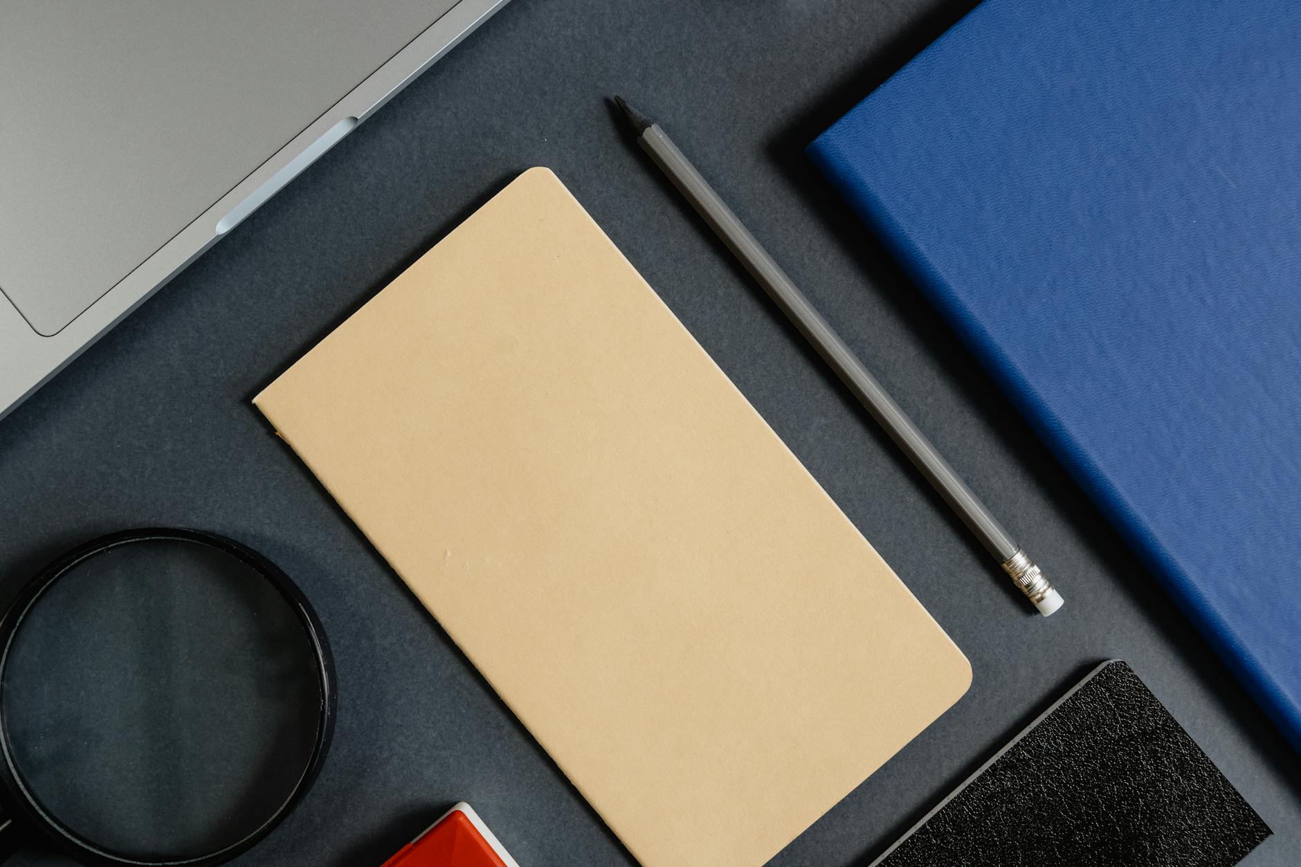 Moody overhead view of dark walnut desk with open laptop showing clean website layout, single warm desk lamp casting amber light across scattered design sketches, editorial still-life photography, warm vermillion and deep charcoal color grade